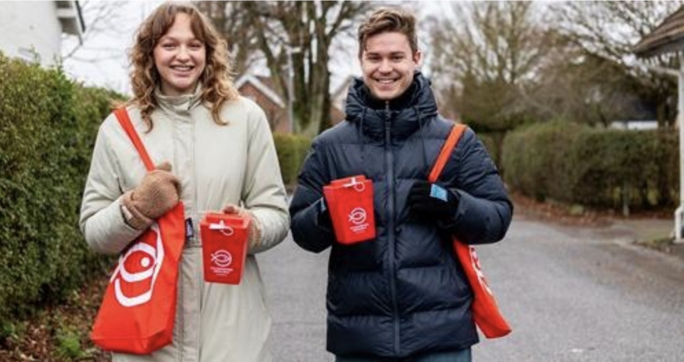 En person lægger en 50-kroneseddel i en rød donationsboks mærket 'Folkekirkens Nødhjælp', som holdes af en anden person iført en mørk jakke.