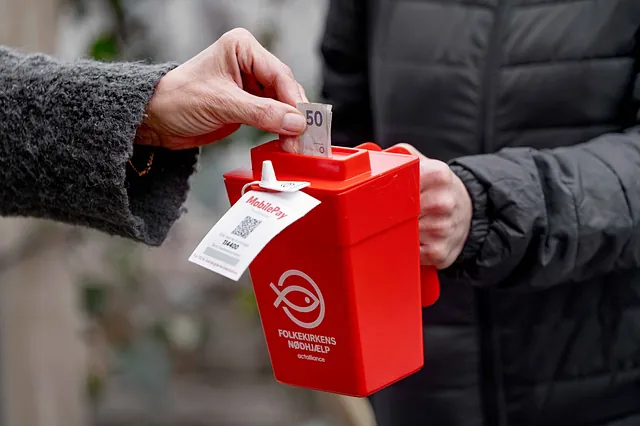 En person lægger en 50-kroneseddel i en rød donationsboks mærket 'Folkekirkens Nødhjælp', som holdes af en anden person iført en mørk jakke.