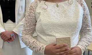 En gruppe unge mennesker klædt i formelt tøj, sandsynligvis til en konfirmationsceremoni, står i kø uden for en kirke på en solrig dag.