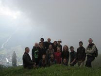 En gruppe mennesker poserer sammen på en græsklædt bakketop med et tåget landskab i baggrunden.