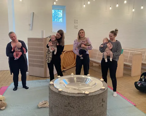 Fire mødre står inde i en kirke med hver deres baby ved en døbefont dekoreret med små tændte stearinlys og med dæmpet belysning i baggrunden.