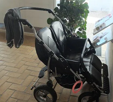 En sort tvillingebarnevogn parkeret indendørs med en grå sweater over håndtaget og et lyserødt stykke legetøj fastgjort til stellet.