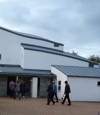 En gruppe mennesker går ind i Tornbjerg Kirke, en hvid kirke med skrånende tag og minimalistisk arkitektur. 