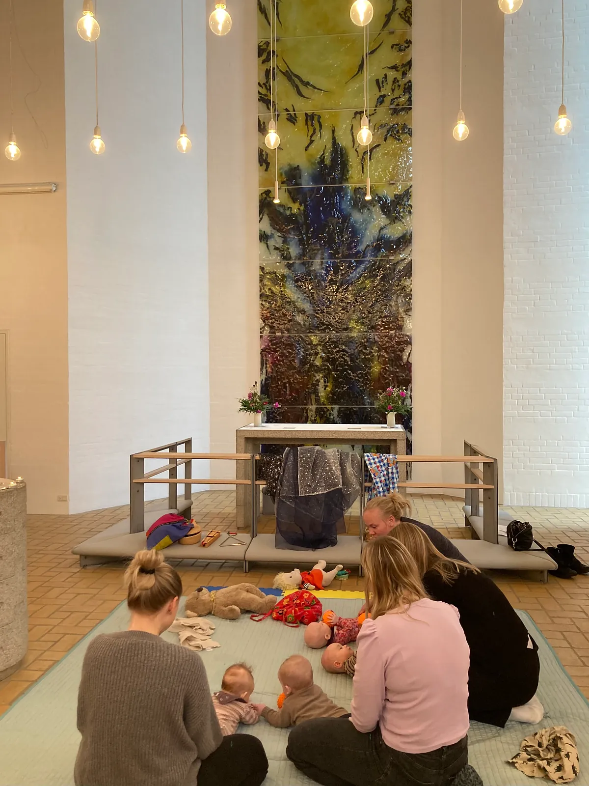 En gruppe mødre sidder på en måtte med deres babyer foran et alter i en kirke, omgivet af dæmpet belysning og et stort, farverigt glaskunstværk.
